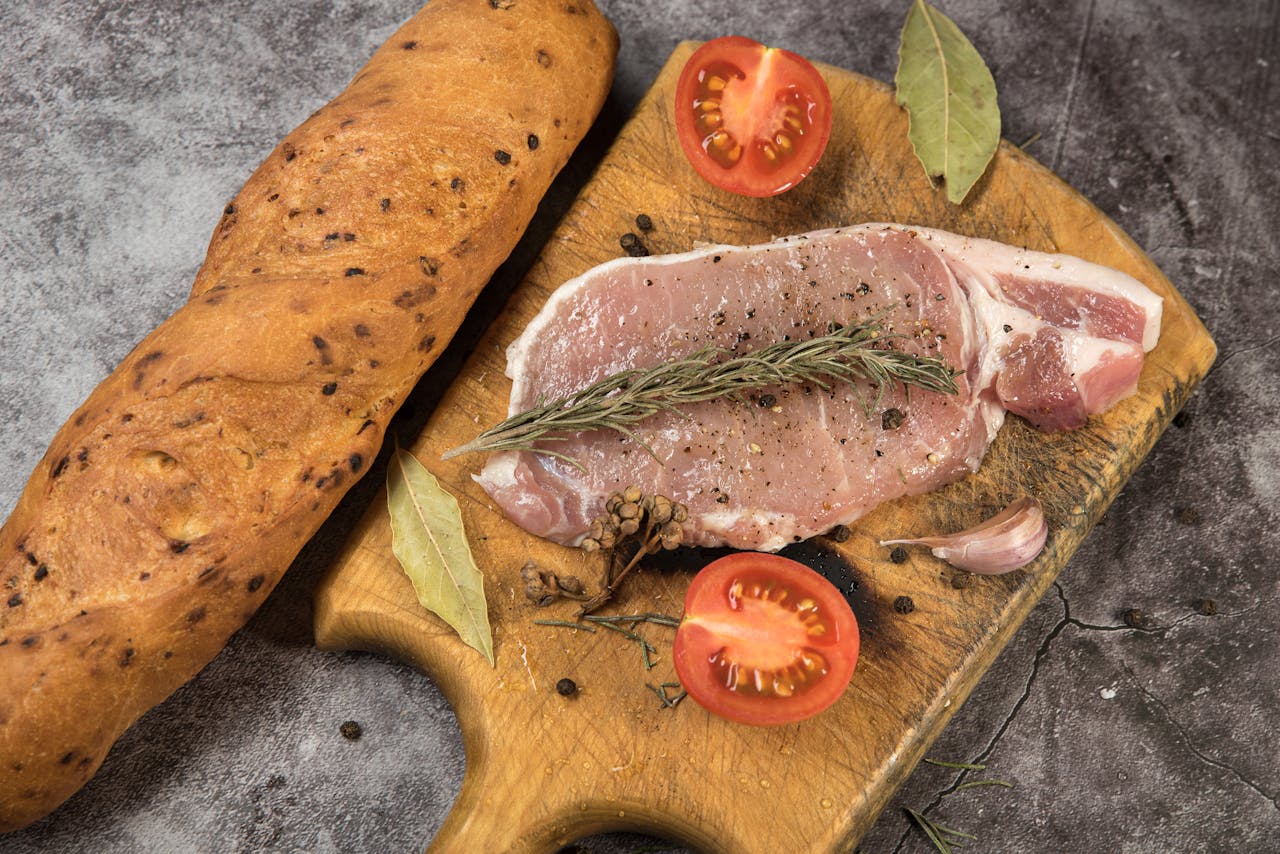 Raw pork chop with rosemary, tomatoes, and bread on wooden board.